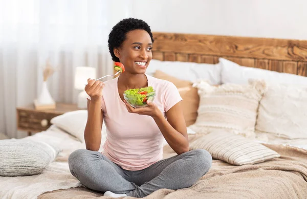 Healthy diet for weight loss concept. Happy black woman eating yummy vegetable salad on bed at home, blank space