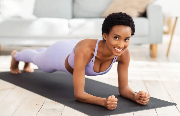 Retrato de comprimento total de mulher negra sorridente em sportswear fazendo prancha de cotovelo no tapete esportivo em casa — Fotografia de Stock