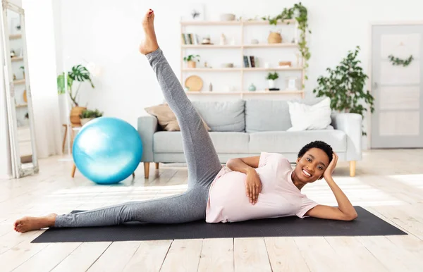 Retrato completo de una mujer negra embarazada haciendo ejercicio sobre una esterilla de yoga, levantando la pierna, sintiéndose saludable en casa — Foto de Stock