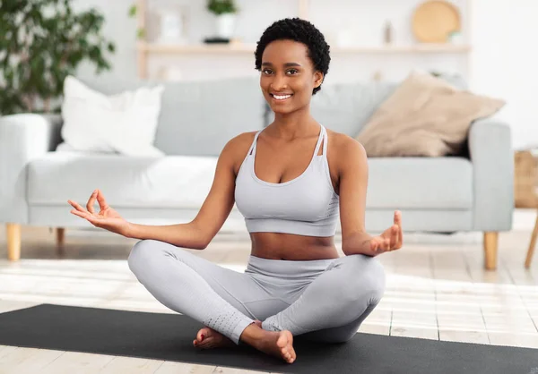 Deportes domésticos durante el concepto de encierro. Mujer negra positiva meditando en pose de loto en casa — Foto de Stock