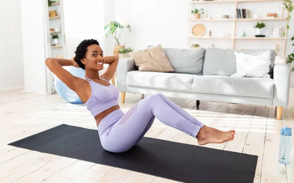 Concepto de entrenamiento en casa. Bonita dama negra en ropa deportiva haciendo ejercicios abdominales en colchoneta de yoga en interiores — Foto de Stock