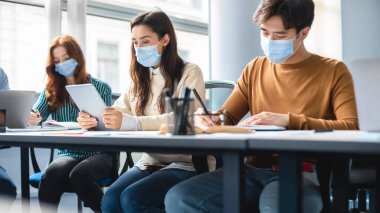 Sınıfta maske takan çeşitli öğrenciler oturuyor.