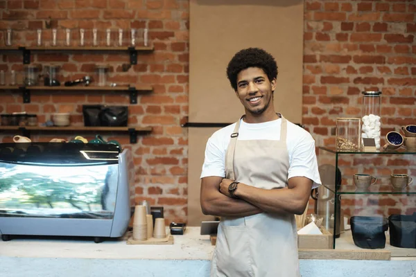 Sorrindo jovem afro-americano em avental, com as mãos cruzadas perto bar contador com equipamento de café — Fotografia de Stock