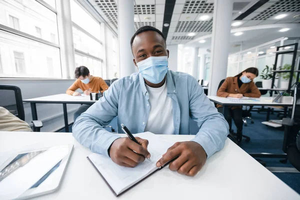 Fekete férfi diák maszkban ül az asztalnál az osztályban — Stock Fotó