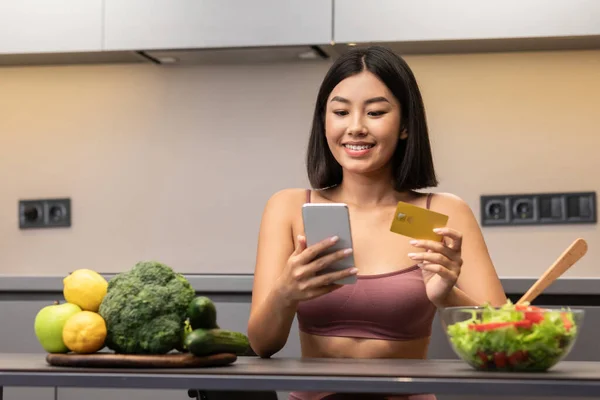 Asiatin mit Telefon kauft Lebensmittel online in der Küche — Stockfoto