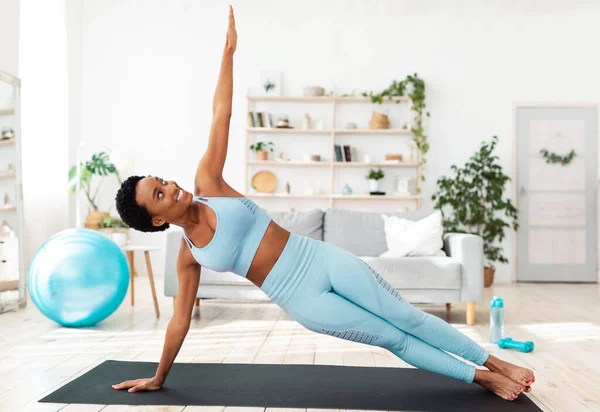 Concepto de fitness casero. Determinada mujer negra ejercitándose en la alfombra deportiva en el interior, haciendo ejercicio de tablón lateral —  Fotos de Stock