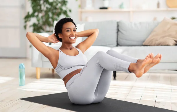 Young black woman with trained body doing abs exercise on yoga mat indoors