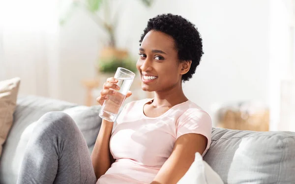 Egészségügyi és méregtelenítő fogalom. Vonzó fiatal nő iszik tiszta vizet üvegből a kanapén otthon — Stock Fotó
