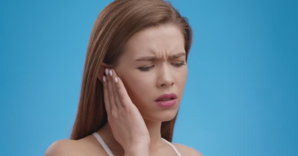 Overstuur jonge vrouw met pijnlijk oor, plotseling gevoel van sterke pijn, gezondheidsprobleem, blauwe achtergrond, close-up — Stockvideo