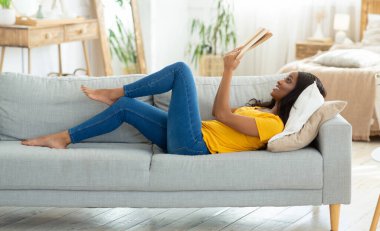 Evde hobilerin olsun. Milenyum siyahi kadın kanepeye uzanmış, kitap açık, yan görüş