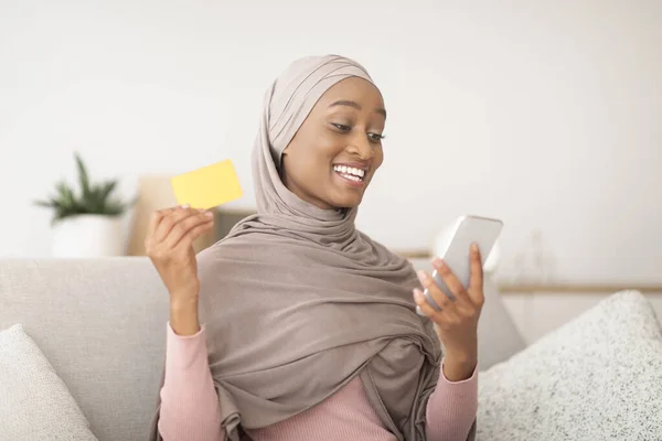 Compras en casa. Señora negra en hijab con tarjeta de crédito y teléfono inteligente, haciendo la compra en línea en el sofá en el interior —  Fotos de Stock