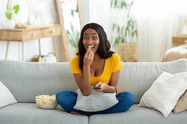 Πλήρες πορτρέτο της νεαρής μαύρης γυναίκας με τηλεχειριστήριο βλέποντας τηλεόραση και τρώγοντας ποπ κορν στον καναπέ στο σπίτι — Φωτογραφία Αρχείου