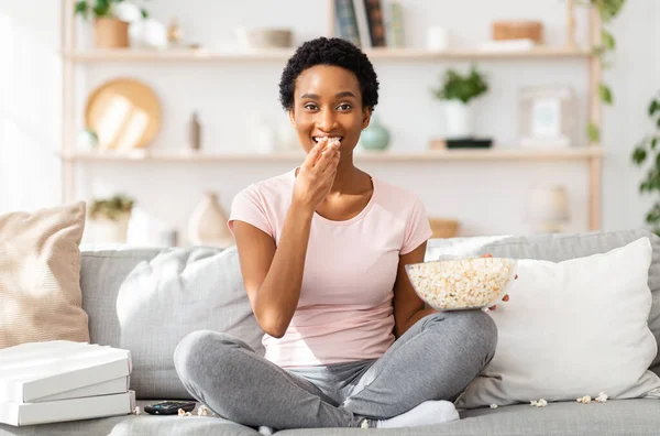 Conceito de comida sem graça. Jovem mulher negra mastigando pipocas na frente da TV no sofá confortável em casa — Fotografia de Stock