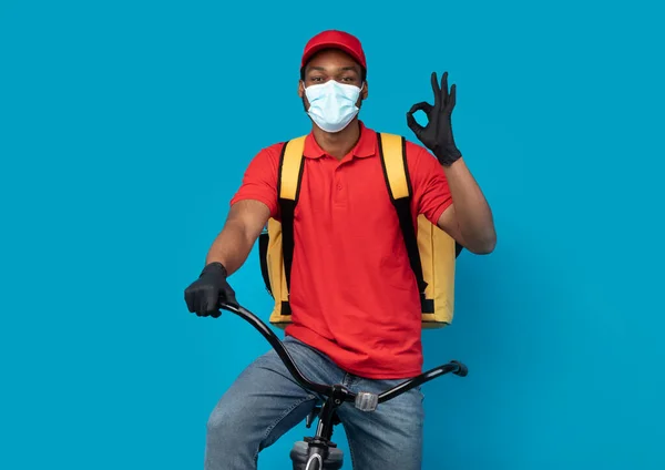 Black delivery man in mask showing okay gesture, riding bike