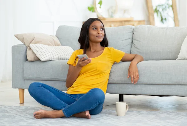 Απομακρυσμένη εκπαίδευση, ιδέα διαδικτυακής εργασίας. Σκέψη μαύρη γυναίκα με κινητό τηλέφωνο κάθεται στο πάτωμα στο σπίτι, αντιγραφή χώρου — Φωτογραφία Αρχείου