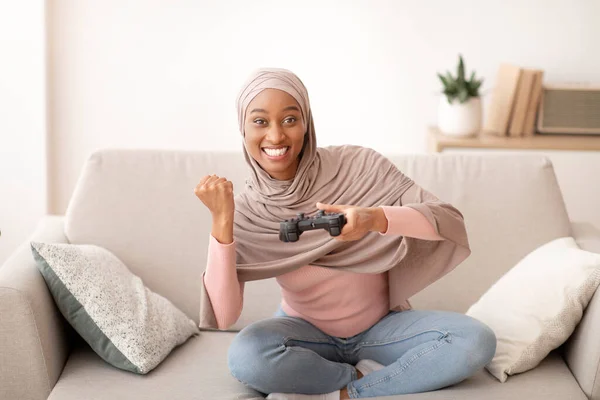 Hétvégi időtöltés. Boldog fekete nő hidzsáb joystick játszik videojátékok beltérben, hogy IGEN gesztus — Stock Fotó