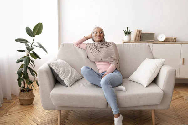 Retrato de comprimento total da mulher negra no hijab descansando no sofá confortável em casa — Fotografia de Stock