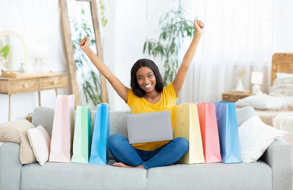 Excited black lady shopping online, sitting on sofa with laptop computer, surrounded by gift bags, indoors