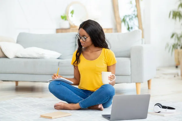 Smart black female student taking notes during online lecture on laptop, studying on floor at home, full length portrait