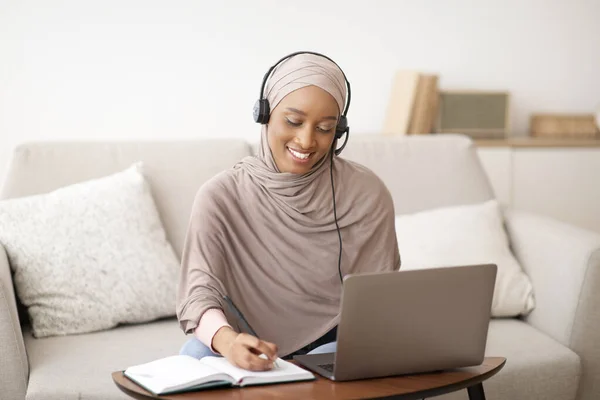 Mulher negra feliz no hijab usando fones de ouvido, assistindo webinar no laptop, tomando notas em casa — Fotografia de Stock
