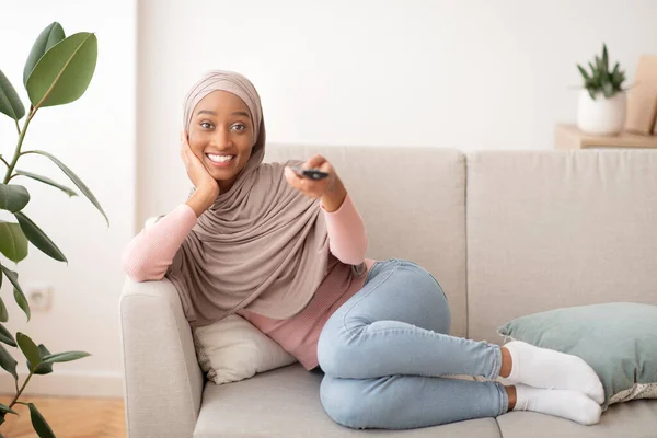 Pozitív fekete nő hidzsábban TV-t néz otthon a kanapén, másolja a helyet — Stock Fotó