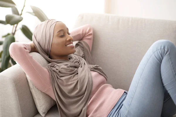 Preciosa dama negra en hijab acostada en el sofá, cerrando los ojos y sonriendo, durmiendo la siesta al mediodía, en el interior — Foto de Stock