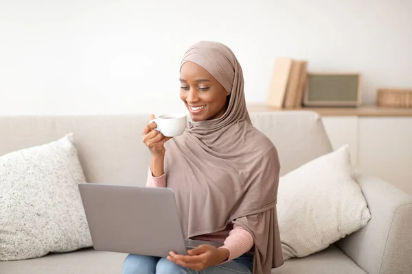 Concetto di vita online. Donna nera in hijab seduta sul divano con computer portatile, bere caffè, lavorare o studiare da casa — Foto Stock