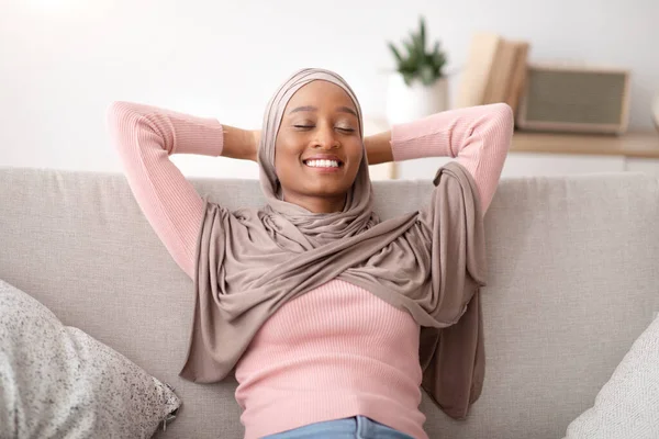 Wanita muslim yang cantik dalam jilbab tradisional bersantai di sofa dengan mata tertutup, di dalam ruangan — Stok Foto