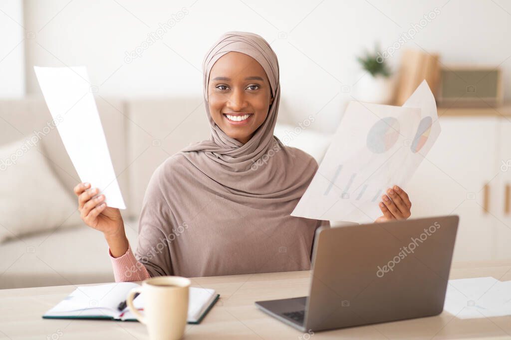 Positive black Muslim woman in hijab working from home, holding documents near laptop computer