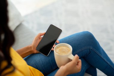 Tanımlanamayan siyahi kadın, beyaz ekranlı akıllı telefon kullanıyor ve evde kahve içiyor, tasarım için maket yapıyor.