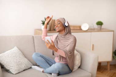 Evde kal. Güzel Müslüman kadın tesettür içinde müzik dinliyor ve evdeki kanepede dans ediyor, boy boy portresi yapıyor.