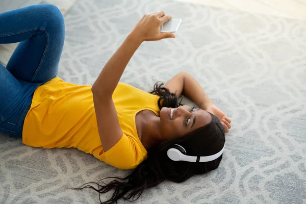 Felice signora afroamericana in cuffia ascoltare musica mentre sdraiato sul pavimento a casa — Foto Stock