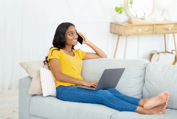 Gioiosa donna afroamericana che comunica con qualcuno sul cellulare e laptop da casa — Foto Stock