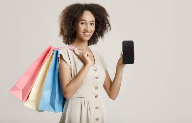 Mutlu Afrikalı Amerikalı genç bayan elinde bir sürü renkli paket tutuyor ve akıllı telefon gösteriyor.