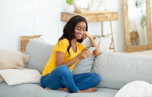 Πλήρες πορτρέτο της νεαρής μαύρης κυρίας που χρησιμοποιεί κινητό τηλέφωνο για online επικοινωνία στον καναπέ στο σπίτι — Φωτογραφία Αρχείου