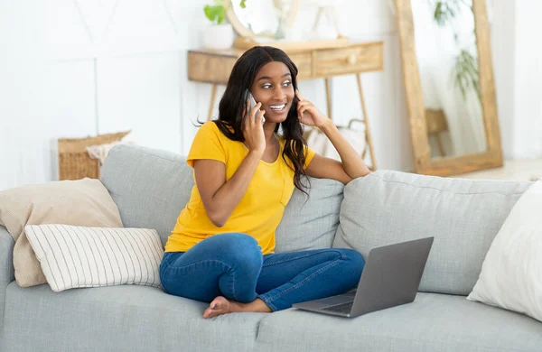 Allegro giovane donna di colore che chatta sullo smartphone e utilizza il computer portatile sul divano di casa — Foto Stock
