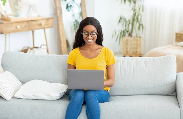 Joyeux dame noire assise sur un canapé confortable avec ordinateur portable et communiquer avec son ami à la maison — Photo