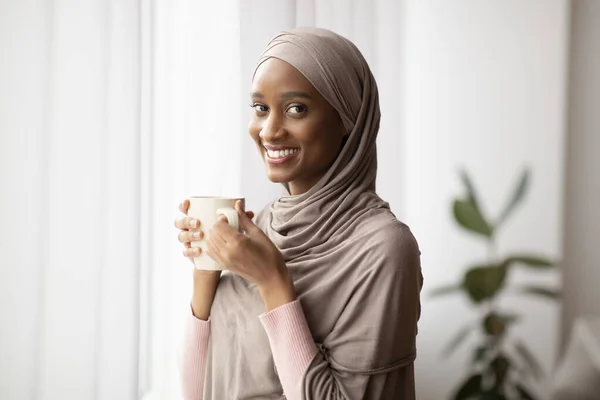 Black Muslim woman in traditional hijab drinking hot aromatic coffee near window at home, having peaceful weekend — Stock Photo, Image