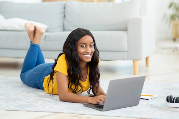 Bella signora nera sdraiata sul pavimento con computer portatile moderno, studiare a distanza o avere riunioni di lavoro, all'interno — Foto Stock