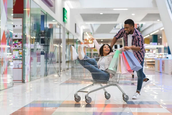 Alegre homem negro puxando carrinho de compras com sua namorada dentro, se divertindo — Fotografia de Stock