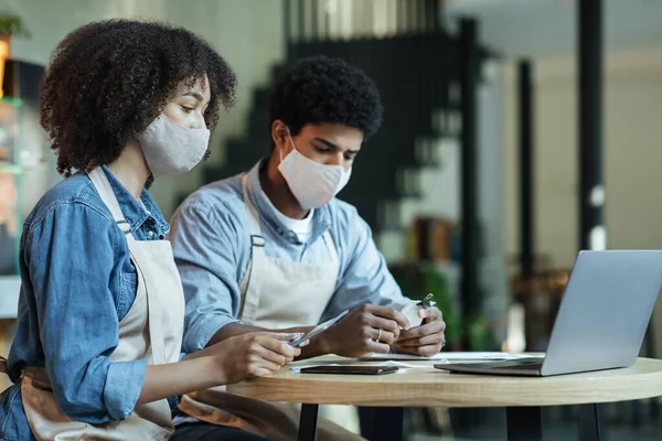 Millennial afro-americano feminino e masculino em aventais e máscaras protetoras sentar à mesa e trabalhar com laptop — Fotografia de Stock