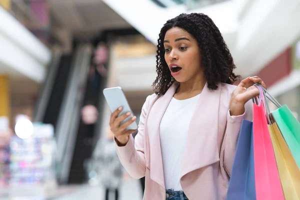 Porträtt av chockad svart kvinna med shoppingväskor och smartphone i köpcentret — Stockfoto