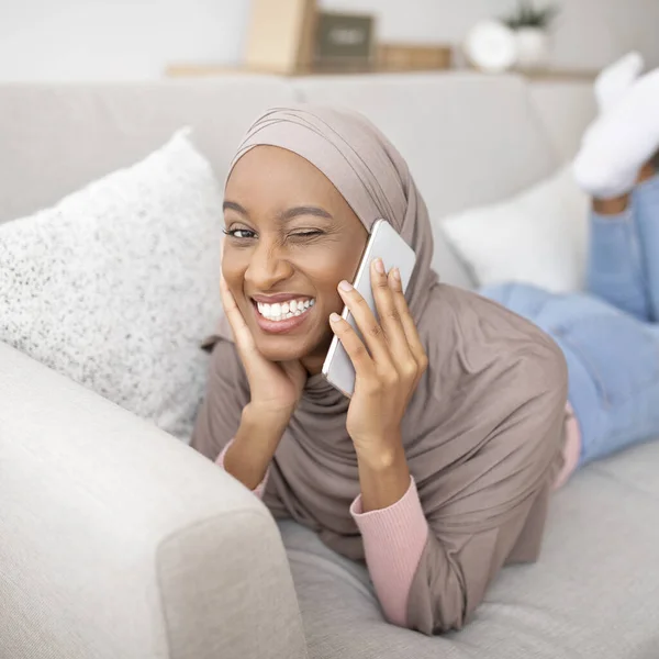 Linda mulher negra no hijab falando no telefone celular e piscando o olho no sofá em casa — Fotografia de Stock