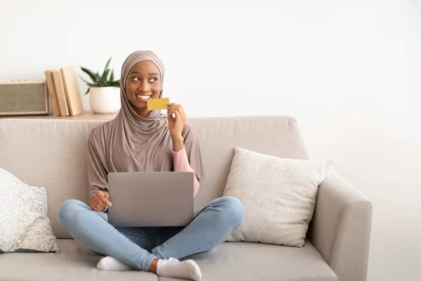 Vidám afro-amerikai hölgy hidzsáb ül kanapén laptop és hitelkártya, vásárlás online otthon, másolás hely — Stock Fotó