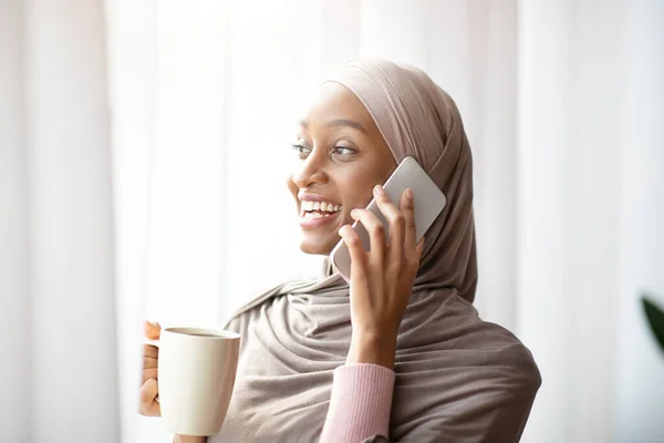 Güzel Müslüman kadın tesettürlü kahve içiyor ve evinin penceresinin yanında telefonla konuşuyor. — Stok fotoğraf