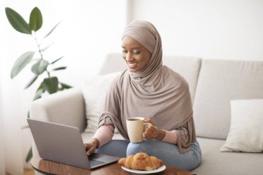Çevrimiçi iş, eğitim, iletişim konsepti. Laptop kullanan ve evde kahve içen tesettürlü mutlu siyah kadın.