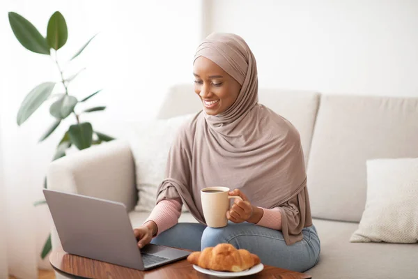 Trabalho online, educação, conceito de comunicação. Senhora preta feliz no hijab usando laptop e beber café em casa — Fotografia de Stock