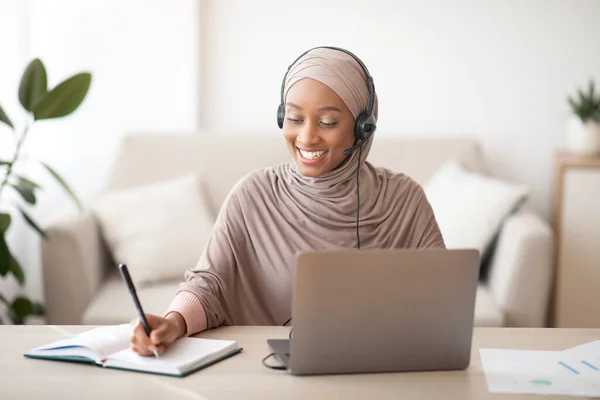 Senhora muçulmana negra em hijab e fone de ouvido tendo videoconferência no laptop, tomando notas durante a aula on-line em casa — Fotografia de Stock