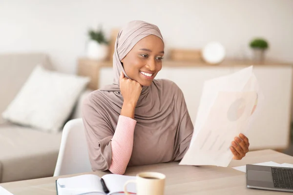 Távmunka koncepció. Pozitív fekete muszlim hölgy hidzsáb tanul dokumentumokat közelében laptop beltéri — Stock Fotó