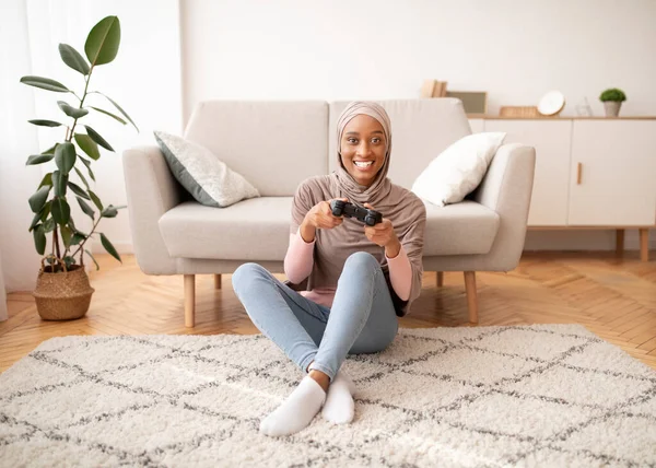 Boldog muszlim nő hidzsábban ül a padlón joystickkal, videojátékozik otthon, teljes hosszúságú portré — Stock Fotó